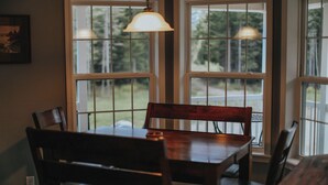 Un comedor con mesa y sillas de madera, ventanas amplias con vistas a zonas verdes y una lámpara colgante.
