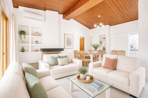 Ein modernes Wohnzimmer mit weißen Sofas, einem Glastisch und einem Essbereich mit Holztischen und einer Tischplatte.