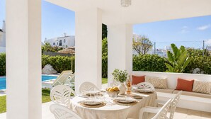 Een terras met een eettafel voor vier personen, witte stoelen, een bank met kussens en uitzicht op een zwembad en groen.