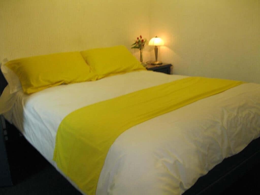 A bed with a white and yellow bedspread, a bedside lamp, and a small vase with flowers.