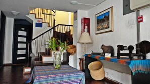 Una habitación con una escalera, una mesa con mantel estampado, una lámpara, una cesta y una figurita de oso.