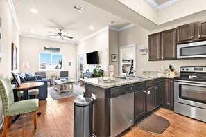 A modern kitchen with stainless steel appliances, a dining area with a table and chairs, and a living room with a sofa and a television. - Alerio E304 (Miramar Beach)