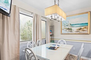 A room with a glass table, white chairs, a painting of a beach scene, a TV, and a lamp. - Blue Surf 19 (Miramar Beach)