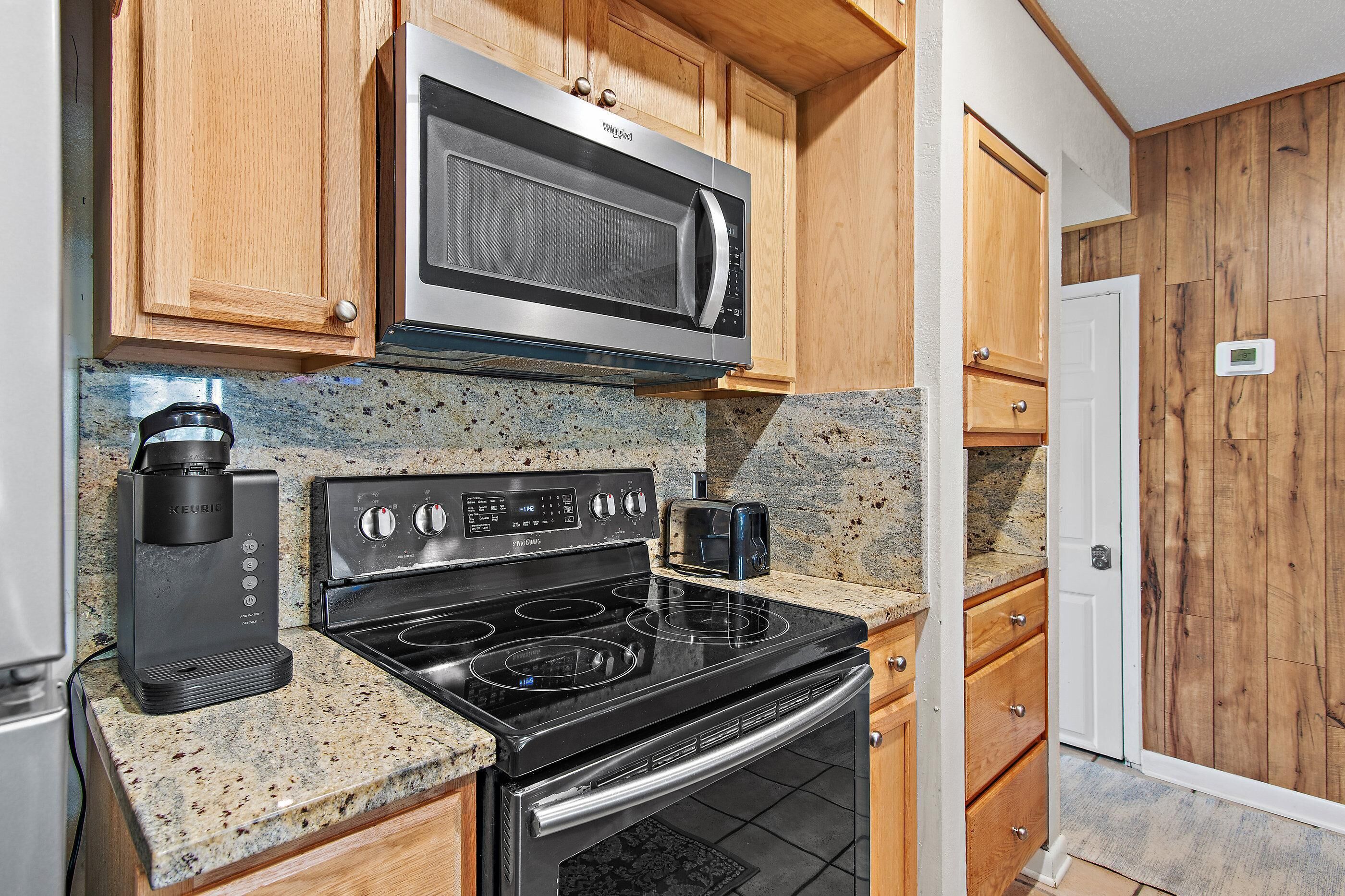 A kitchen with a microwave, coffee maker, stove, oven, granite countertop, wooden cabinets, and a door.