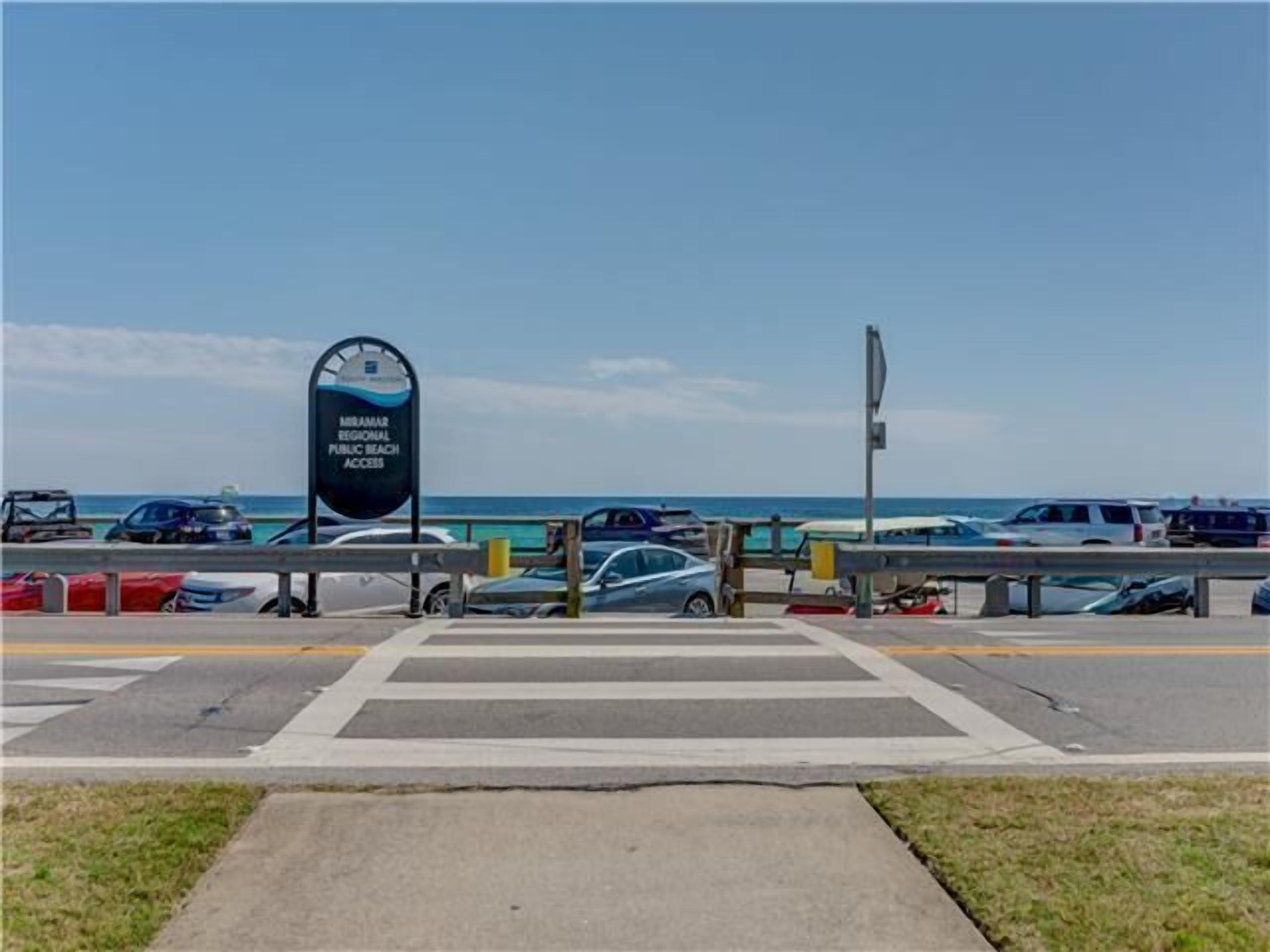 En parkeringsplats med en skylt som visar vägen till den allmänna stranden Miramar Regional Public Beach, ett övergångsställe och fordon som är parkerade längs vägkanten.