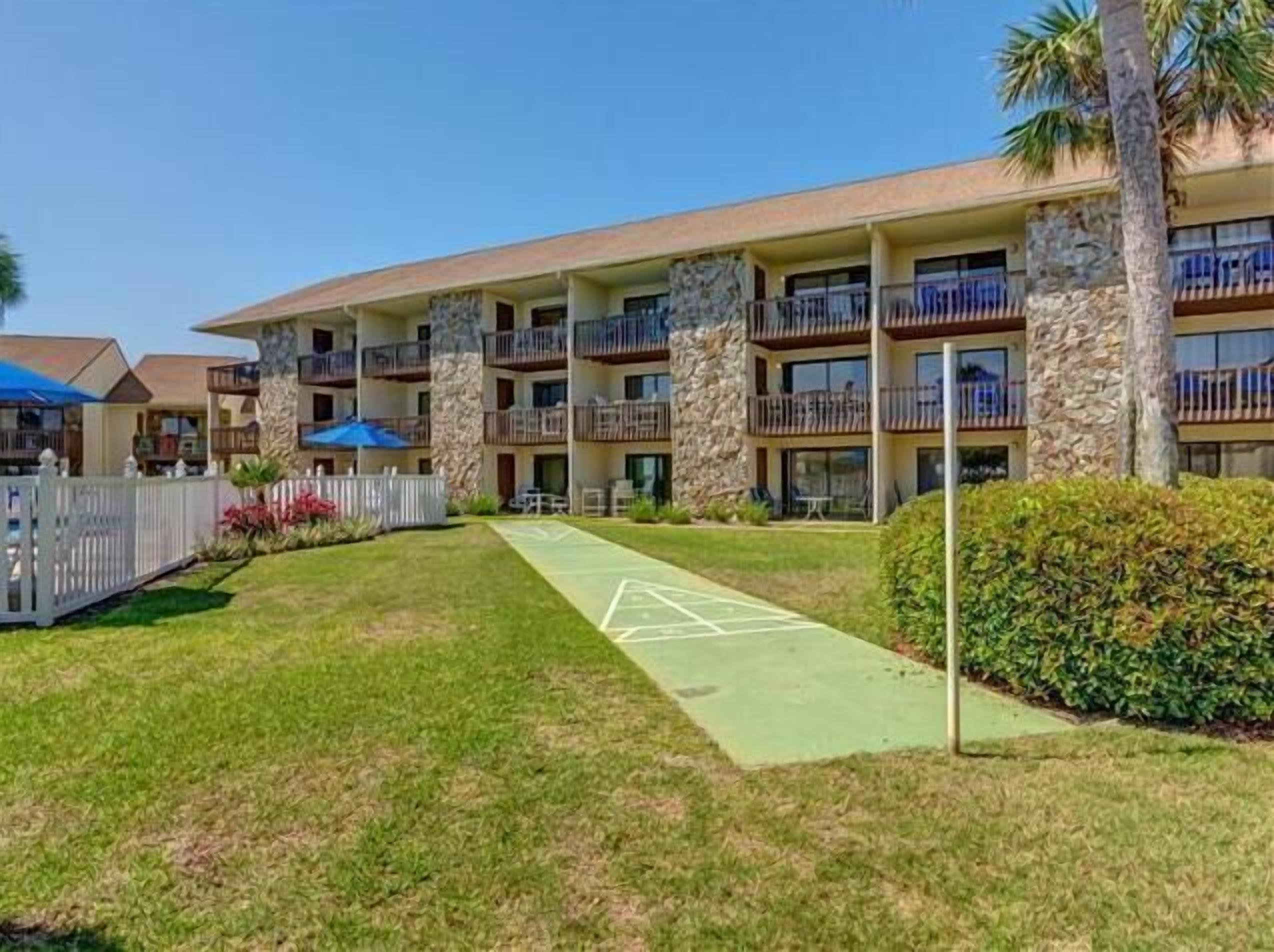 Un hotel de varios pisos con balcones, un patio con césped verde y una cerca blanca de piquete.