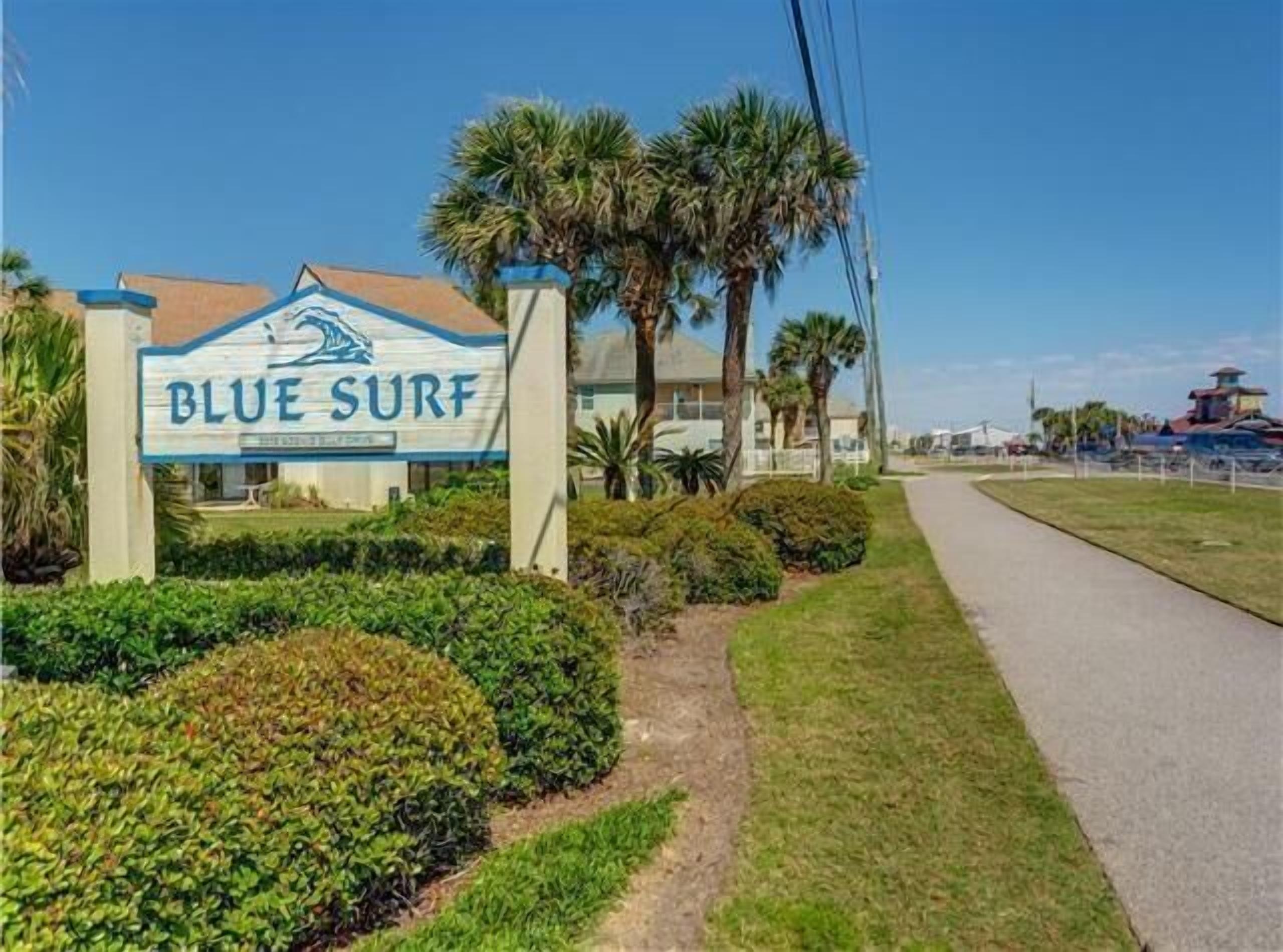 Une enseigne indiquant « Blue Surf », avec un bâtiment en arrière-plan, entouré de verdure et sous un ciel dégagé.