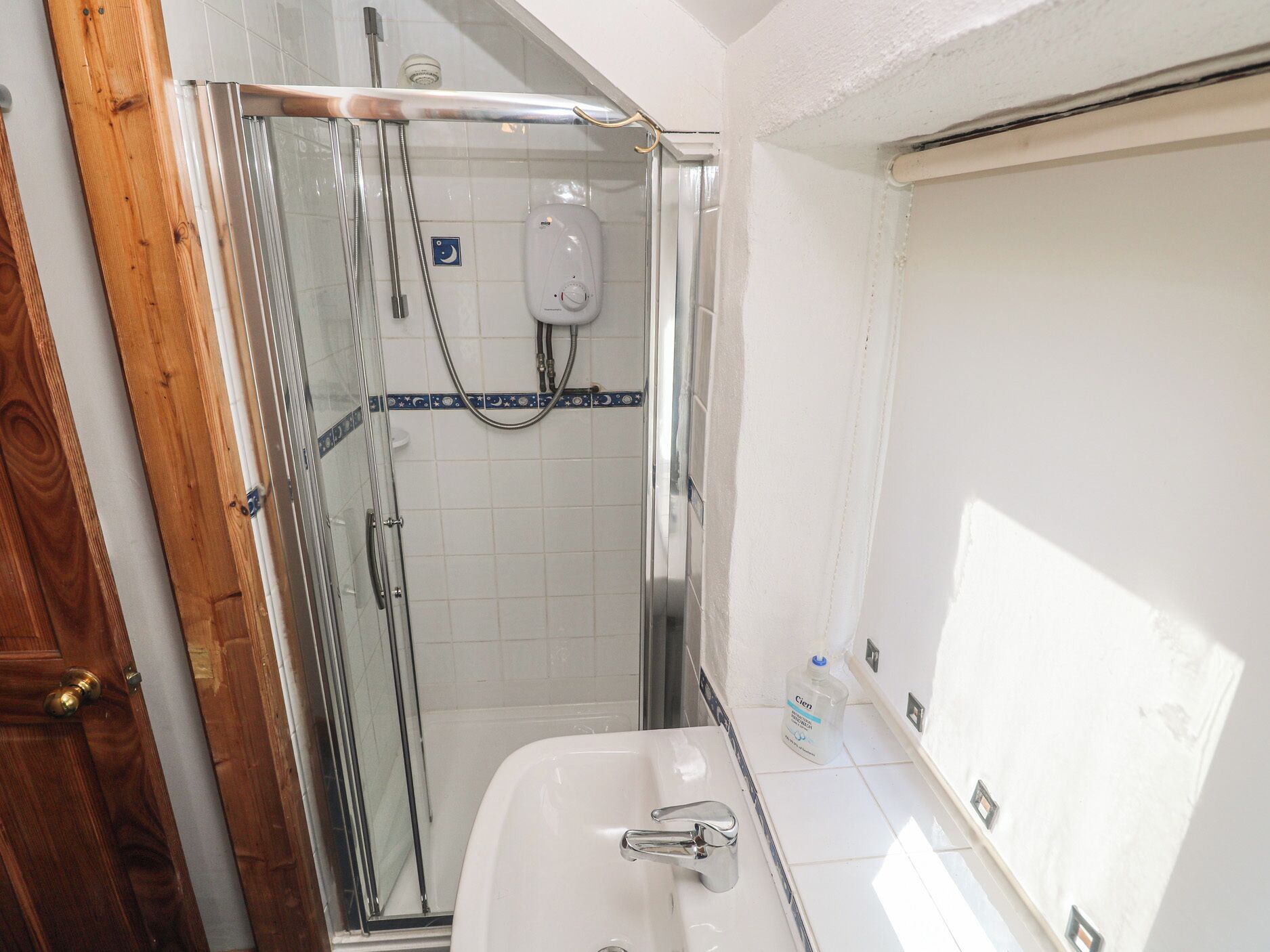 Een badkamer met een douchecabine van glas, een witte wastafel en een fles handzeep op het aanrecht.
