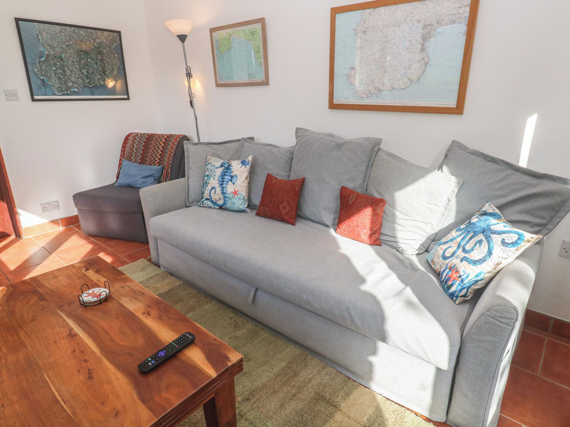 Ein Wohnzimmer mit grauem Sofa, einem Holztisch und einer Fernbedienung darauf. An der Wand hängen drei gerahmte Karten.