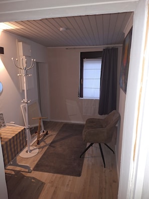 A room with a wooden floor, a chair, a desk with a chair, a coat rack, a window with curtains, and a ceiling with exposed beams.