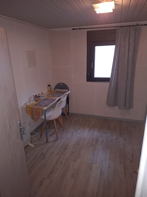 Una habitación pequeña con piso de madera, una mesa con sillas, un ventanal con cortinas y una lámpara fijada en la pared.