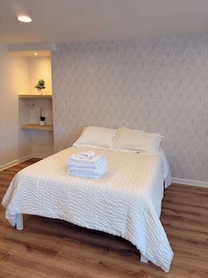 A neatly made bed with a textured white duvet, a small bedside table with a potted plant, and a patterned wall.