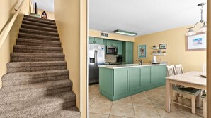 Un escalier mène à une cuisine dotée de meubles de couleur verte, d’un réfrigérateur et d’un coin repas avec une table et des chaises.
