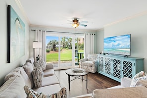 A living room with a sofa, a coffee table, a TV, and a view of a garden through large windows. - Ciboney 1001 (Miramar Beach)