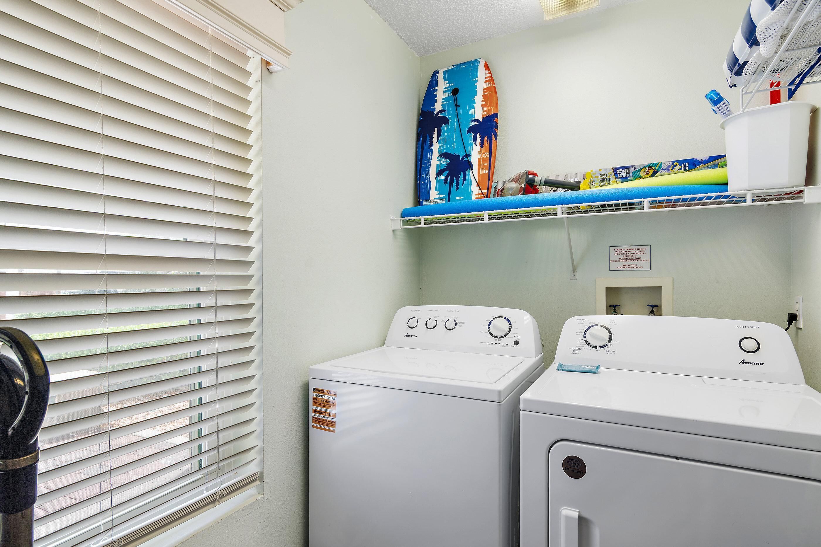 A laundry area with two white washers and dryers, a window with blinds, and a shelf with various items.
