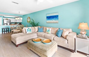Ein Wohnzimmer mit einem beigefarbenen Ecksofa, einem Couchtisch aus Holz mit zwei Schalen, einer Küche im Hintergrund und einer Türkiswand mit einem Strandbild.