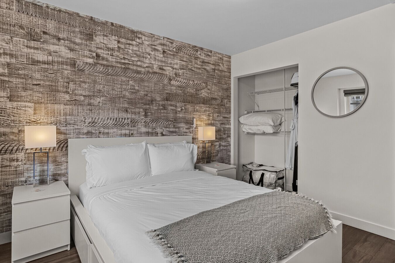 Un dormitorio moderno con una pared texturizada, una cama con ropa de cama blanca, una lámpara de noche, un espejo circular y un armario con ropa colgada.