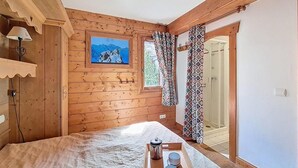 Een houten kamer met een bed, een schilderij, een raam met gordijnen met een patroon, en een douchegedeelte zichtbaar door een open deur.