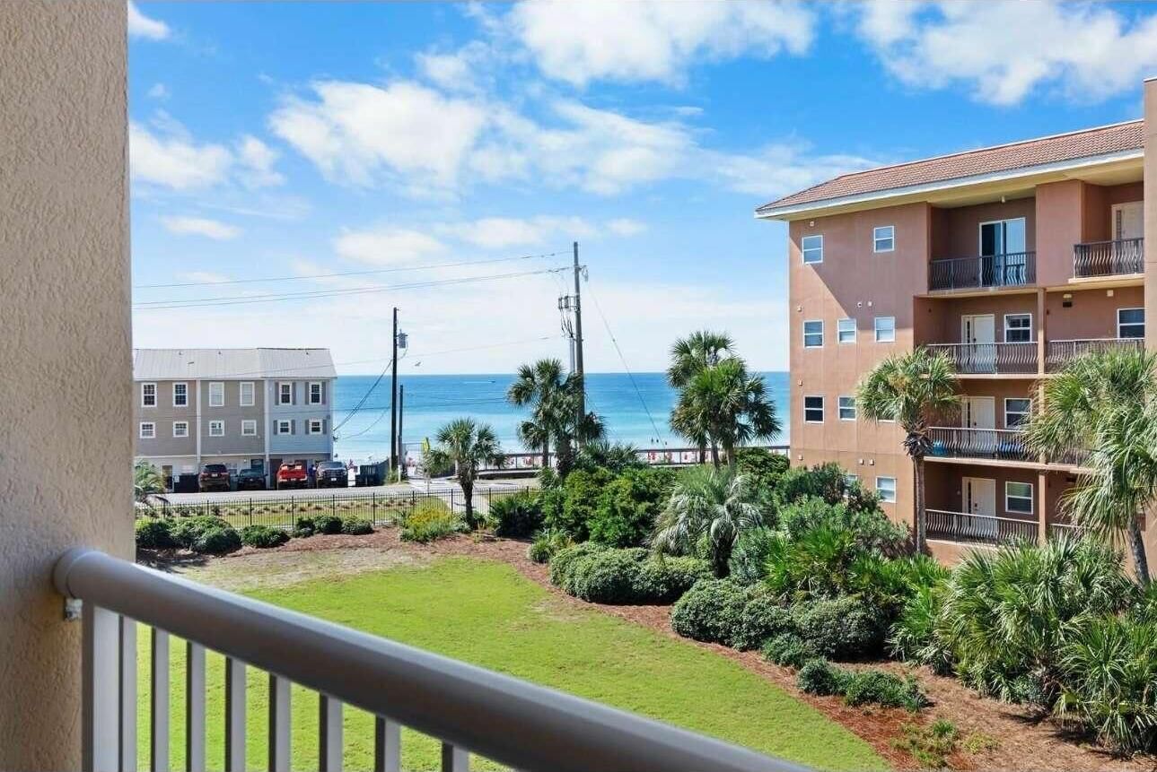 A balcony with a view of the ocean, a parking lot, and a building with multiple floors and balconies.