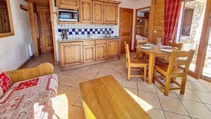 Cozinha acolhedora com armários de madeira, micro-ondas e painel de azulejos. Há uma área de jantar com mesa e cadeiras de madeira, e um sofá com capa estampada.