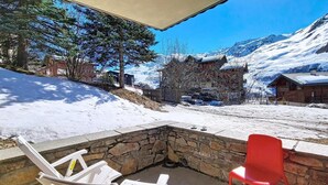 Área de pátio com lareira de pedra, cadeiras brancas e vermelhas, e vista para montanhas cobertas de neve.