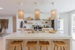Een moderne keuken met een centraal kookeiland, barkrukken, hangende lampen en een koelkast.