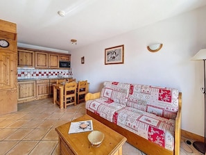 A cozy living area with a sofa, wooden dining table, and chairs, a kitchen with wooden cabinets, and a tiled backsplash. - 1st floor, swimming pool, balcony, tv, ski locker, 32m², Les Menuires (Les Menuires)
