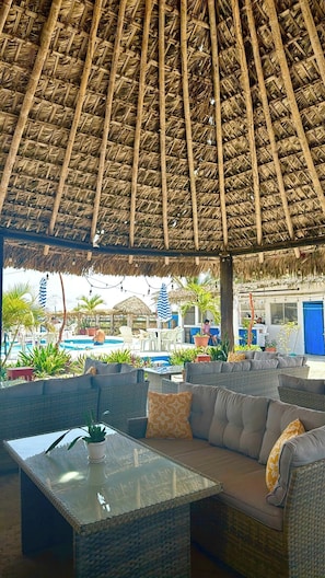 A covered outdoor seating area with wicker furniture and a glass-top table, overlooking a pool and a building with a blue door. - PACHA BEACH (Tuxpan)