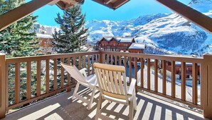Una terraza de madera con dos sillas blancas, vista a montañas nevadas y un edificio con techo inclinado.