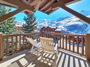 Eine Holzterrasse mit zwei weißen Stühlen, Blick auf schneebedeckte Berge und ein Gebäude mit schrägem Dach.