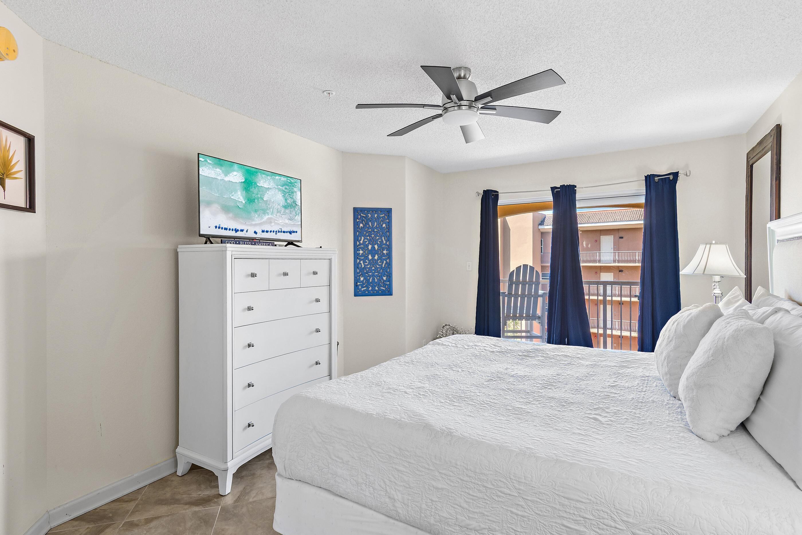 Un dormitorio con cama, ventilador de techo, televisor sobre una cómoda, balcón con cortinas y vistas al exterior.