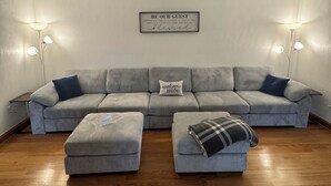 Ein Wohnzimmer mit grauem Sofa, zwei Poufs, einem Wandzeichen und zwei Stehlampen.