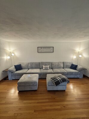 Ein Wohnzimmer mit grauem Sofa, zwei Poufs, einem Wandzeichen und zwei Stehlampen.