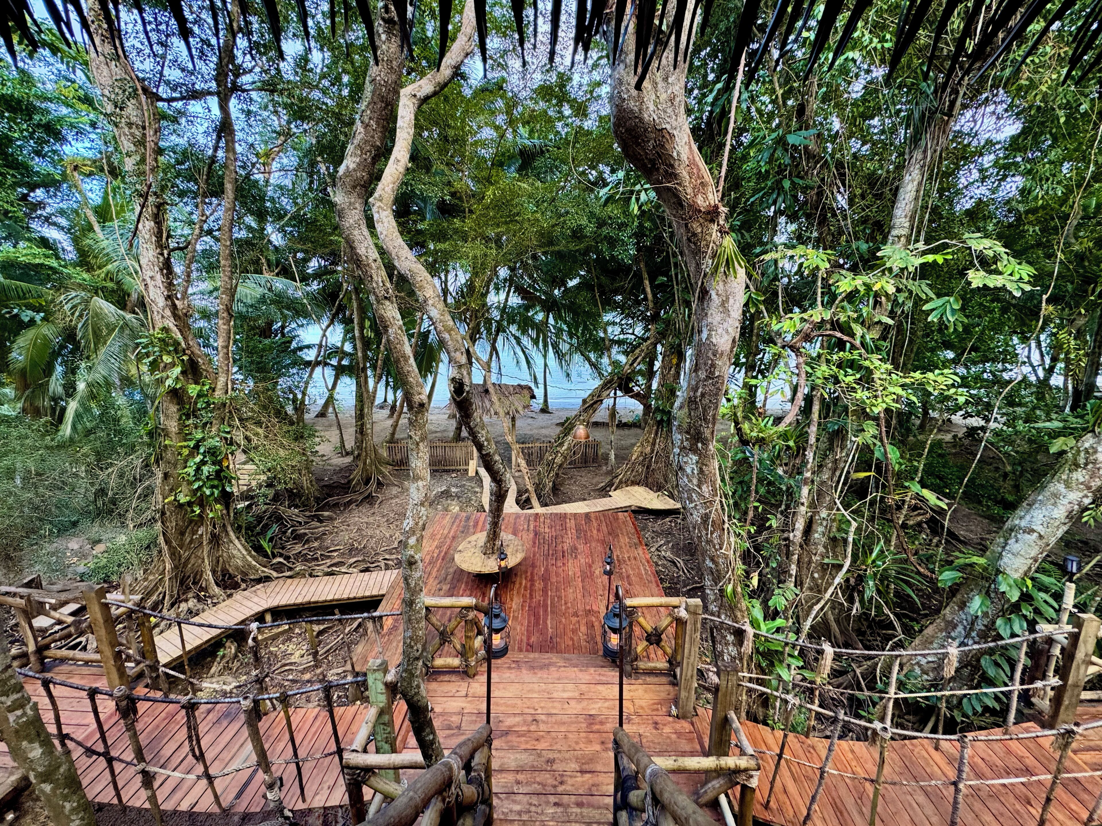 A wooden deck surrounded by trees, leading to a beach area with a thatched-roof structure.