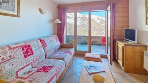 A living room with a patterned sofa, a television on a wooden cabinet, and a view through a glass door. - 100m from ski slopes, floor -1, swimming pool, terrace, tv, ski locker, 32m², Les Menuires (Les Menuires)