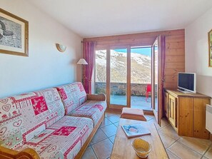 Ein Wohnzimmer mit einem gemusterten Sofa, einem Fernseher auf einer Holzkommode und Blick durch eine Glastür.