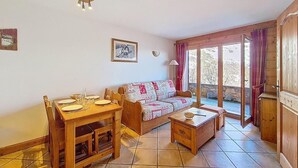 A cozy living room with a wooden dining table, a sofa, and a small coffee table. There is a window with curtains and a view of trees outside. - 100m from ski slopes, floor -1, swimming pool, terrace, tv, ski locker, 32m², Les Menuires (Les Menuires)