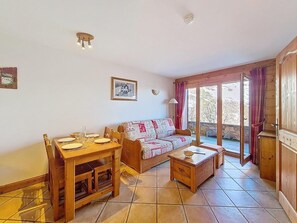 Ein gemütliches Wohnzimmer mit einem Holzesstisch, einer Couch und einem kleinen Couchtisch. Ein Fenster mit Vorhängen bietet Blick auf Bäume.
