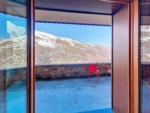 Ein Balkon mit Steinmauer, einem roten Stuhl und Blick auf verschneite Berge.