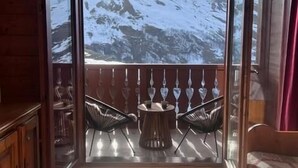 Una habitación con decoración de madera, un balcón con vistas a montañas nevadas, una mesa redonda con sillas y una cama con cortina roja.