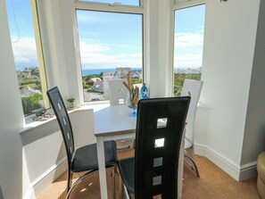 A room with a large window offering a view of the sea and houses, a table with chairs, and a vase with decorative items. - Ewyn Y Don (Holyhead)