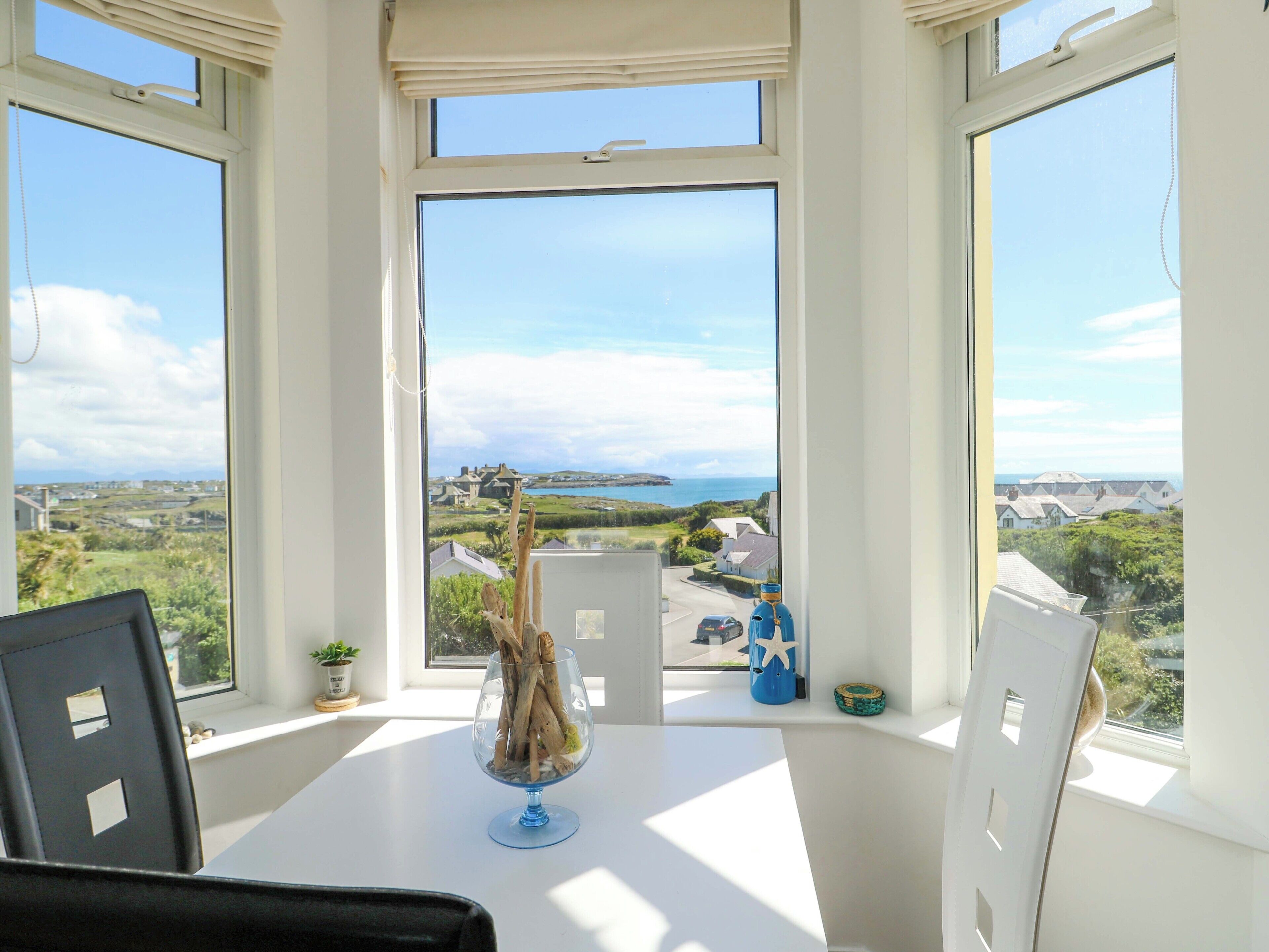 Una habitación con una mesa, sillas, un ventanal con vista a un paisaje costero y un jarrón de cristal decorativo con ramas.