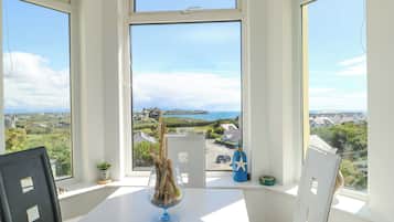 Una habitación con una mesa, sillas, un ventanal con vista a un paisaje costero y un jarrón de cristal decorativo con ramas.