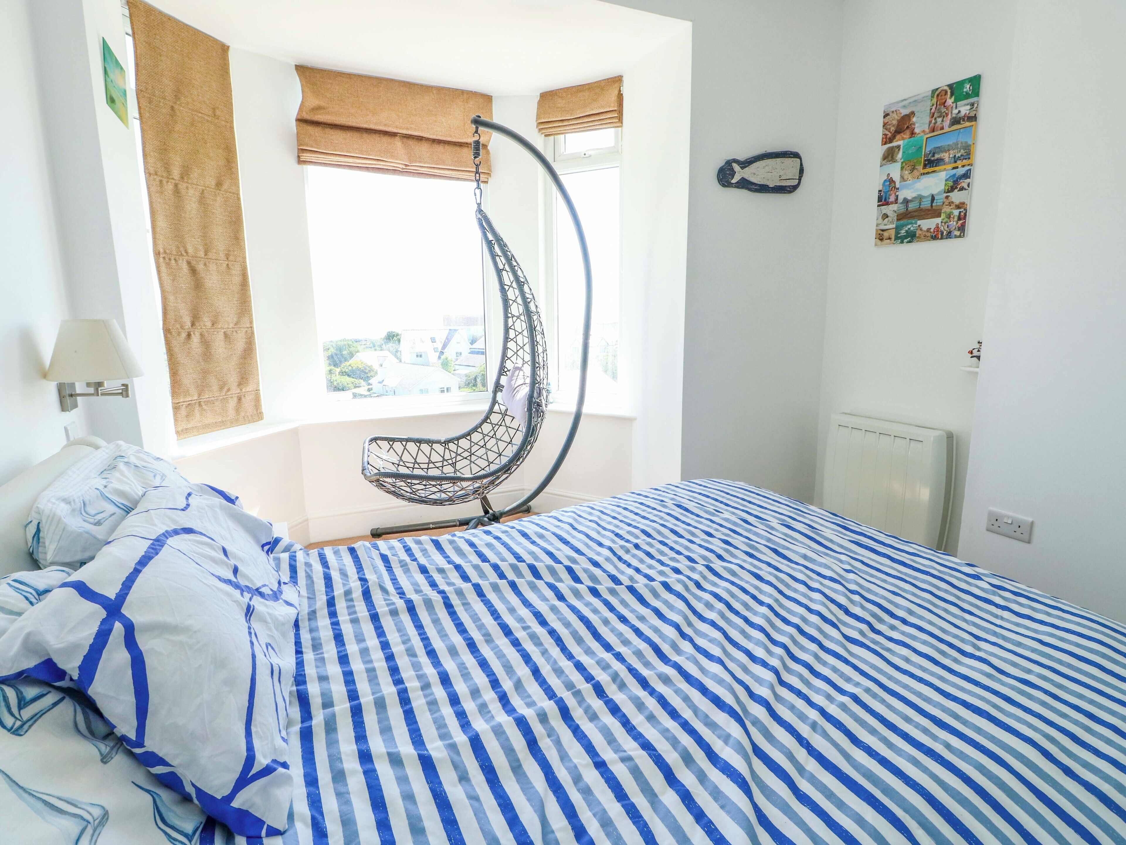 A bedroom with a hanging chair, a bed with striped bedding, a window with a view, and a wall with a fish decoration.
