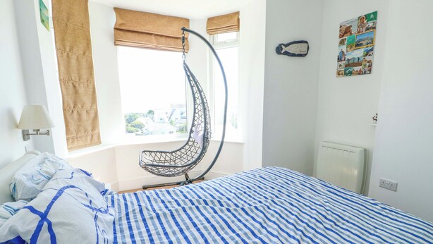 A bedroom with a hanging chair, a bed with striped bedding, a window with a view, and a wall with a fish decoration. - Ewyn Y Don (Holyhead)