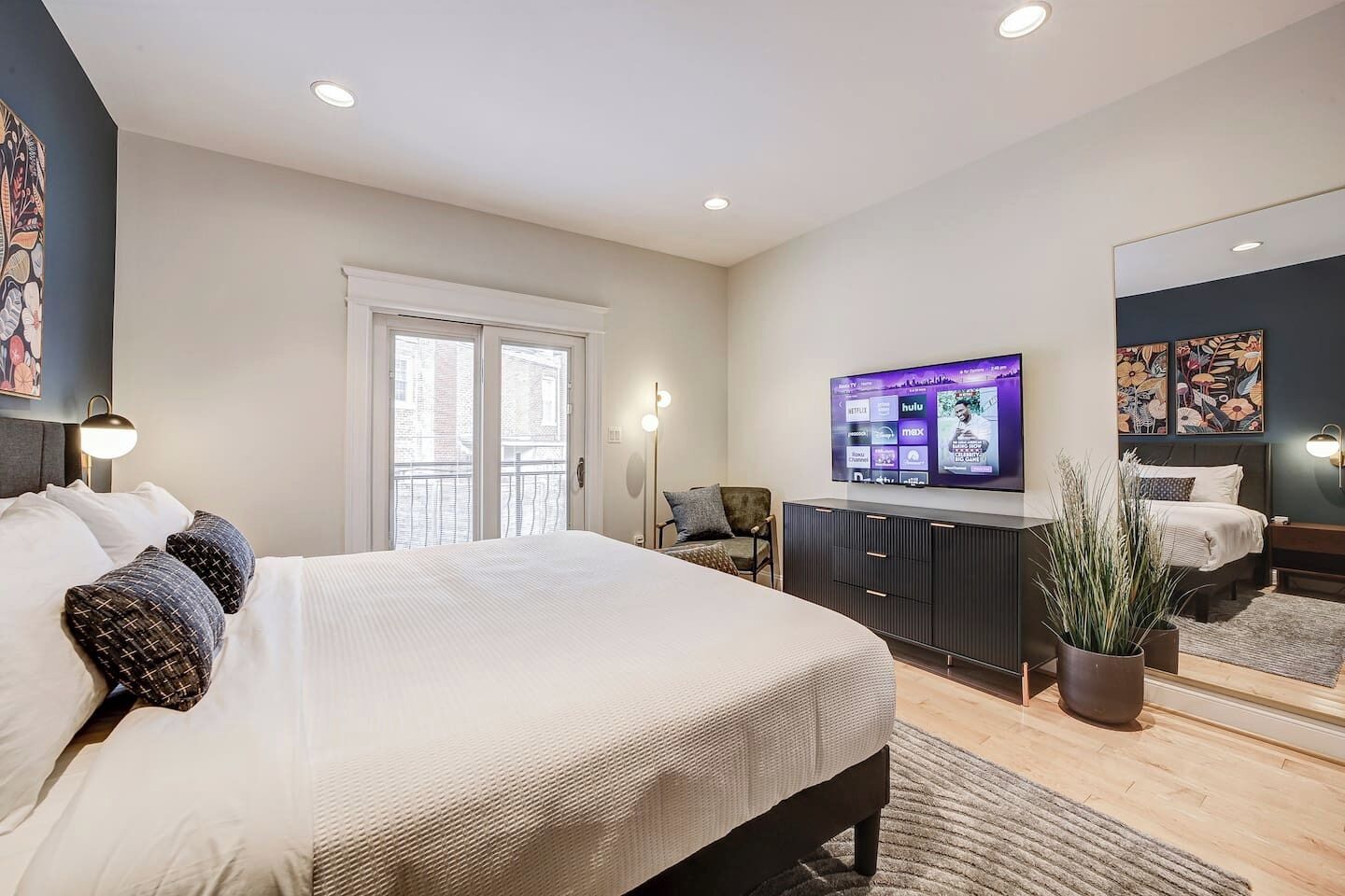 A modern bedroom with a large bed, a TV mounted on the wall, a balcony, and a potted plant.