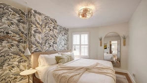 A bedroom with a large bed, a wall-mounted mirror, a ceiling light fixture, and a window with blinds. - Big and Beautiful 5BR Triplex in Shaw/Bloomingdale (Washington)