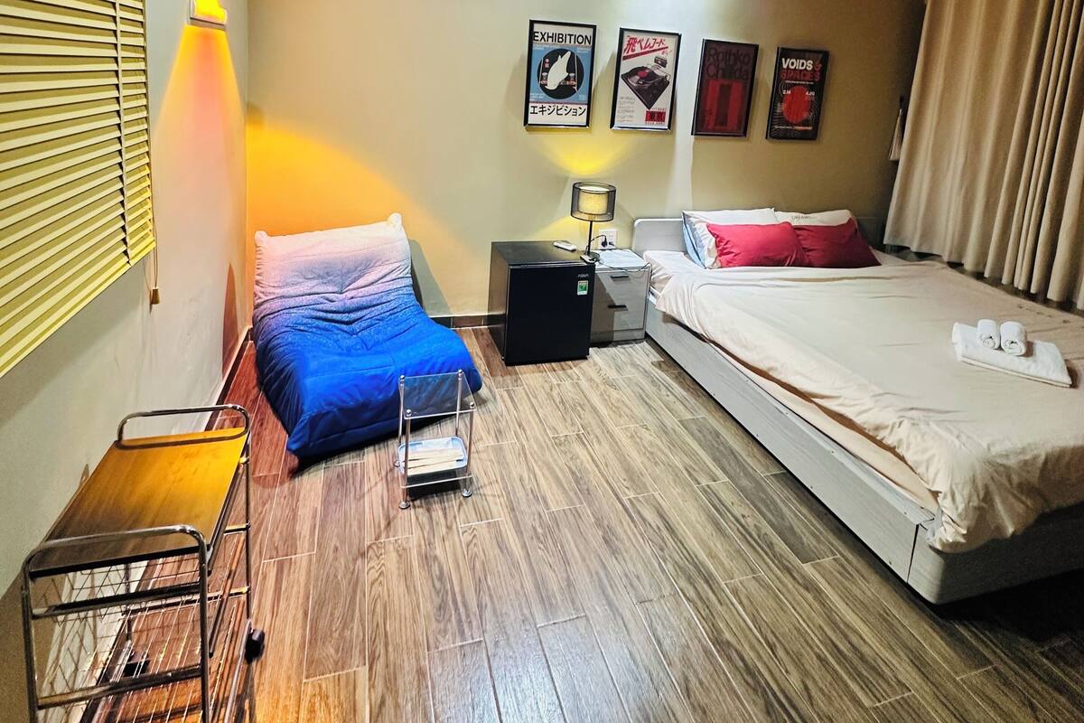 A hotel room with a bed, a desk, a chair, a small table, and a wall-mounted shelf.