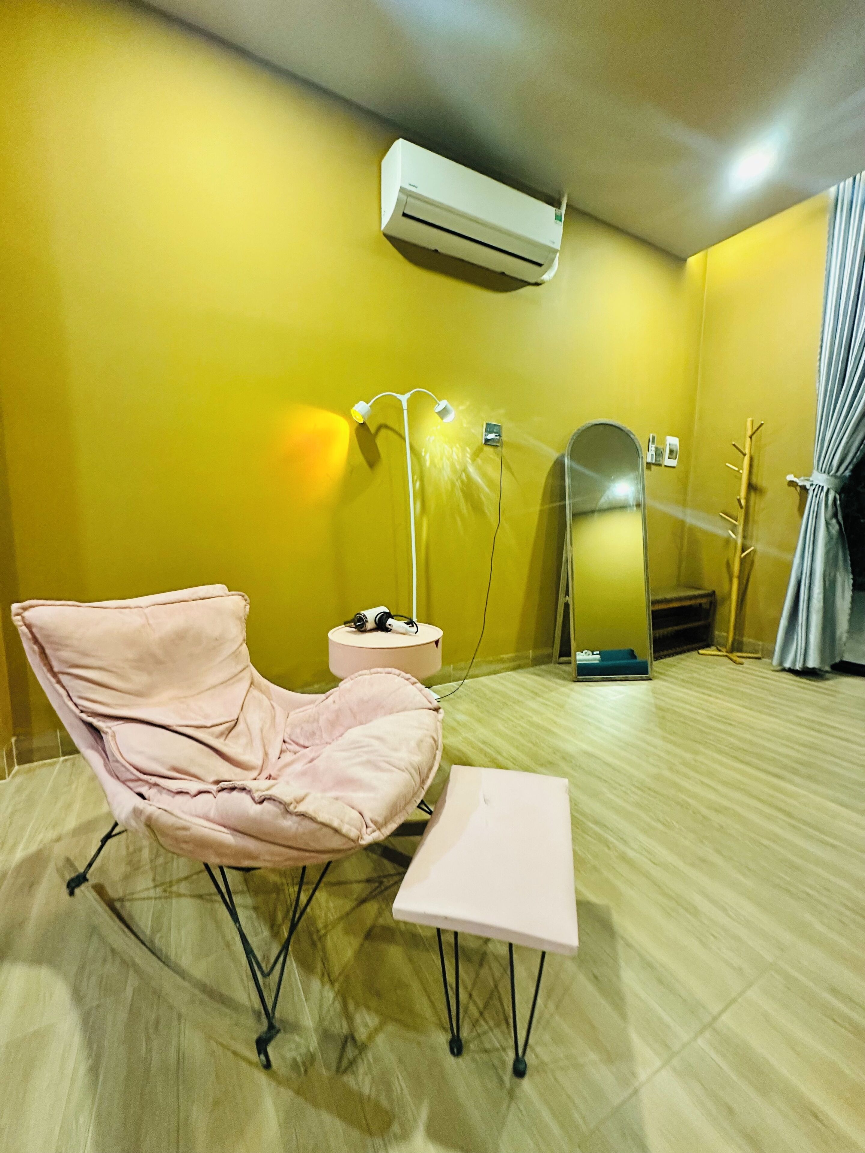 A room with a yellow wall, a white air conditioner, a pink chair, a small table, a mirror, and a floor lamp.