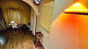 Una habitación con piso de madera, una cama con colcha azul y rosa, una mesita de noche con lámpara, una ventana con persianas y una lámpara de pared.